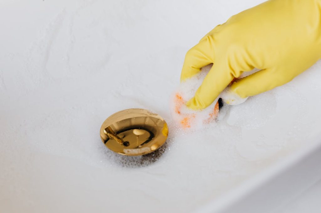 person-in-glove-using-sponge-and-detergent-for-washing-bathroom-sink-4239115 Close-up of a hand in a glove cleaning a foam-covered gold drain in a sink.